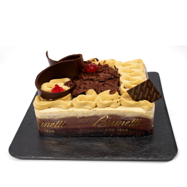 A rectangular brunetti cake with chocolate and vanilla cream swirls, garnished with cherries, chocolate shavings, and a chocolate ribbon, on a slate board.