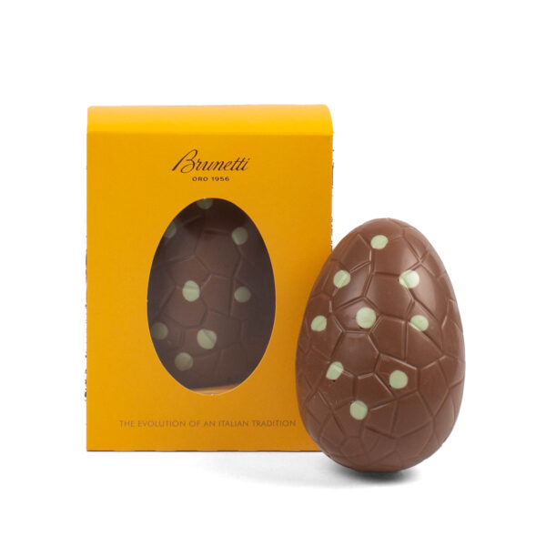A brunetti chocolate easter egg with white dots, standing next to its yellow packaging that features a window showing the product.