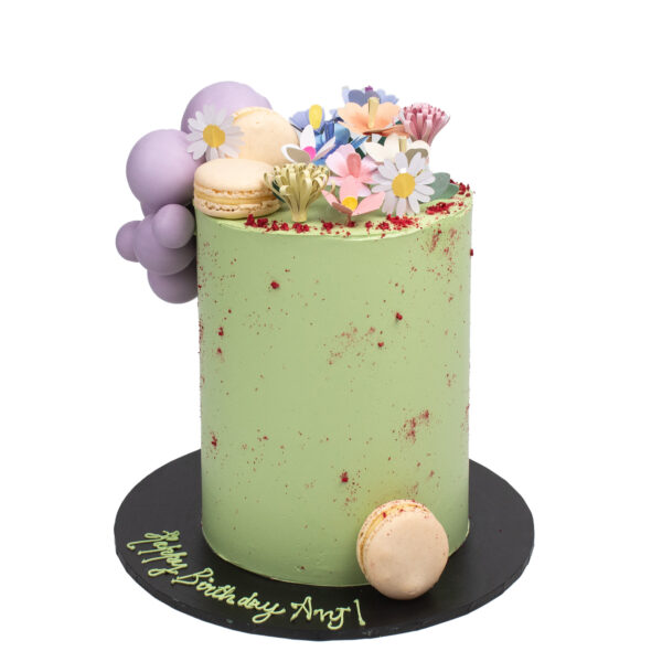A tall green birthday cake decorated with colorful edible flowers, macarons, and a "happy birthday amy" inscription on its base.