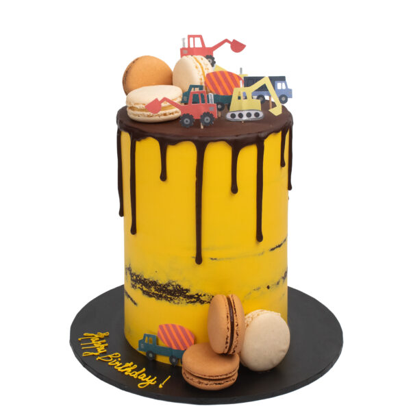 A yellow birthday cake with chocolate drips, topped with macarons and toy construction vehicles, with a "happy birthday" message.