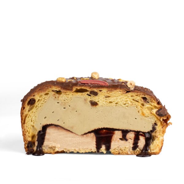 A cross-section of a stuffed panettone with layers of ice cream and chocolate sauce, topped with nuts, against a white background.