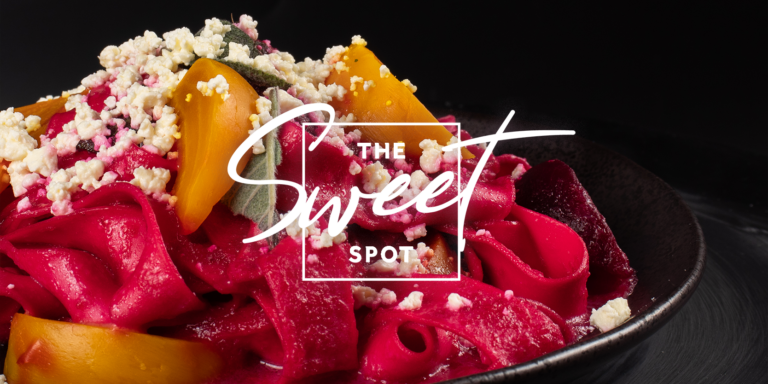 Black plate containing colorful beetroot pasta with yellow bell peppers and crumbled cheese, labeled "the sweet spot" in a decorative white font.