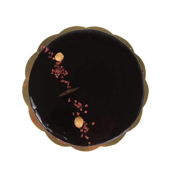 Top view of a dark chocolate glazed Vegan Pistacchiola gelato cake garnished with nuts, dried berries, and a cinnamon stick, placed on a golden cake board against a white background.