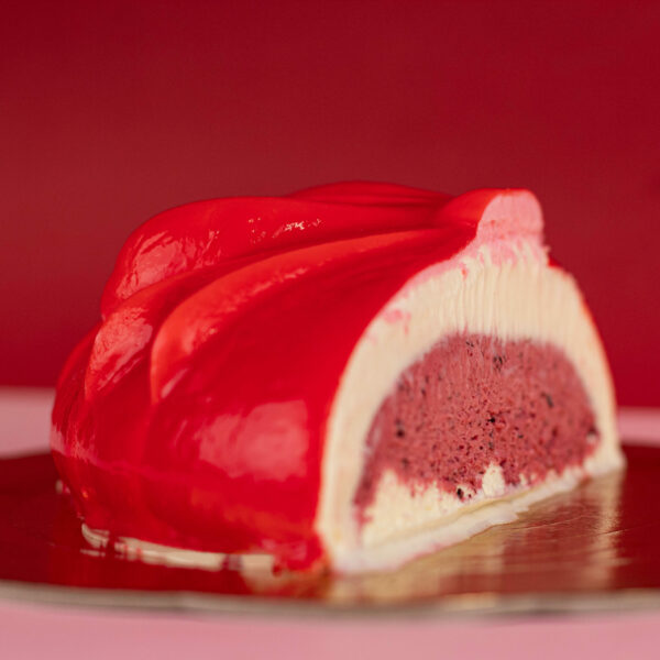 A vibrant, red glazed dessert with a creamy outer layer and a pink, berry-filled center, presented on a gold plate against a red background.